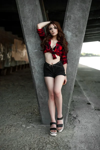 Menina bonita hipster posando e sorrindo perto de fundo da parede urbana em camisa xadrez vermelho, shorts, ao ar livre retrato de verão . — Fotografia de Stock