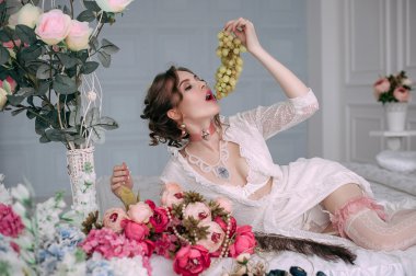 Beyaz dantel elbise, Oda beyaz yatakta oturuyor ve üzüm, yemek güzel genç seksi kadın çiçeklerle süslenmiş. Mükemmel makyaj. Güzellik moda. Kirpik. Stüdyo düzeltisiz atış. 