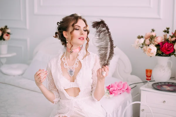 Beautiful young sexy woman sitting on white bed, wearing white lace dress, hair decorated with flowers. Perfect makeup. Beauty fashion. Eyelashes. Studio retouched shot.