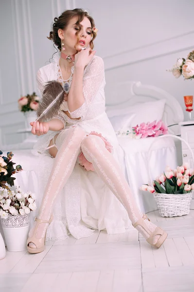 Belle jeune femme sexy assise sur un lit blanc, portant une robe en dentelle blanche, des cheveux décorés de fleurs. Maquillage parfait. Mode beauté. Des cils. Studio retouché . — Photo