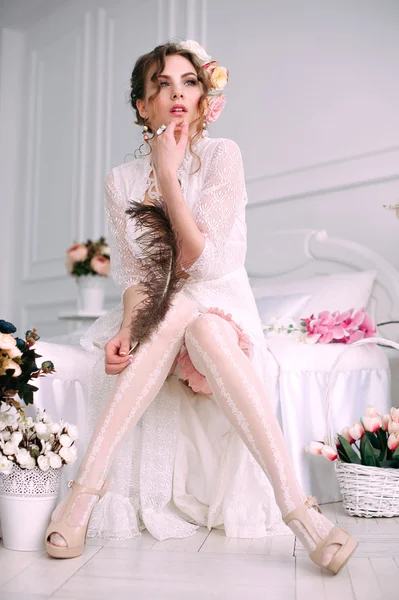 Mulher sexy jovem bonita sentada na cama branca, vestindo vestido de renda branca, cabelo decorado com flores. Maquiagem perfeita. Moda de beleza. Cílios. Estúdio retocado tiro . — Fotografia de Stock
