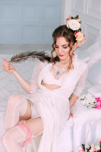 Vacker ung sexig kvinna sitter på vit säng, klädd i vit spetsklänning, hår dekorerad med blommor. Perfekt makeup. Skönhet mode. Ögonfransar. Studio retuscheras skott. — Stockfoto