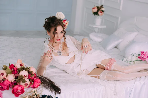 Όμορφη νεαρή σέξι γυναίκα κάθεται στο λευκό κρεβάτι, φορώντας λευκή δαντέλα φόρεμα, μαλλιά, διακοσμημένο με λουλούδια. Τέλειο μακιγιάζ. Ομορφιά μόδα. Βλεφαρίδες. Studio retouched βολή. — Φωτογραφία Αρχείου