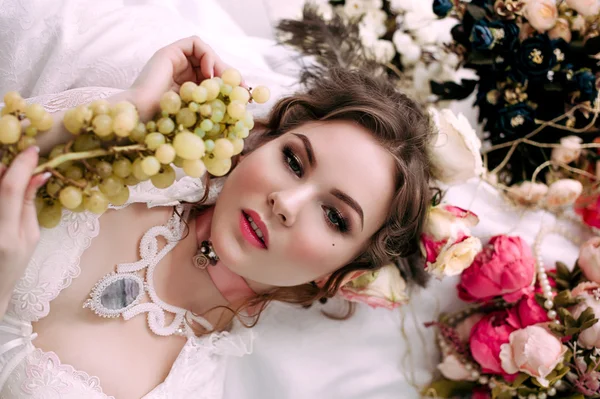 Красивая молодая сексуальная женщина сидит на белой кровати и ест виноград, одетая в белое кружевное платье, комната украшена цветами. Идеальный макияж. Красота. Ресницы. Ретушированный снимок . — стоковое фото