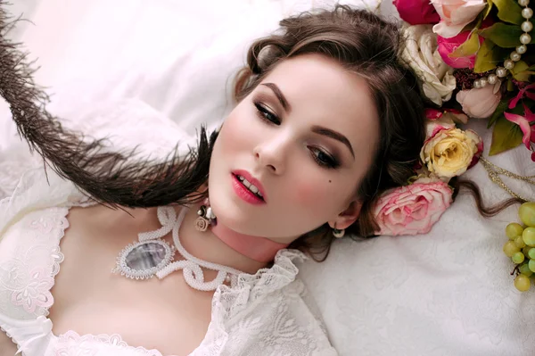 아름 다운 젊은 섹시 한 여자 흰 침대에 앉아서 먹는 포도, 룸, 화이트 레이스 드레스를 입고 꽃으로 장식 되어 있습니다. 완벽 한 메이크업입니다. 뷰티 패션입니다. 속눈썹입니다. Retouched 스튜디오 샷. — 스톡 사진