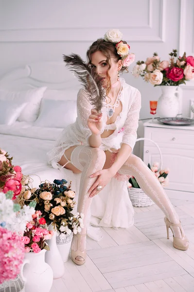 Beautiful young sexy woman sitting on white bed, wearing white lace dress, hair decorated with flowers. Perfect makeup. Beauty fashion. Eyelashes. Studio retouched shot.