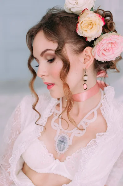 Belle jeune femme sexy assise sur un lit blanc, portant une robe en dentelle blanche, des cheveux décorés de fleurs. Maquillage parfait. Mode beauté. Des cils. Studio retouché . — Photo