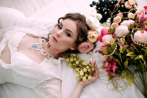 Beautiful young sexy woman sitting on white bed and eating grapes, wearing white lace dress, room decorated with flowers. Perfect makeup. Beauty fashion. Eyelashes. Studio retouched shot.