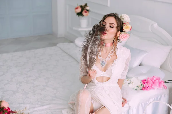 Hermosa mujer sexy joven sentada en la cama blanca, con vestido de encaje blanco, cabello decorado con flores. Maquillaje perfecto. Moda de belleza. Pestañas. Estudio retocado tiro . — Foto de Stock