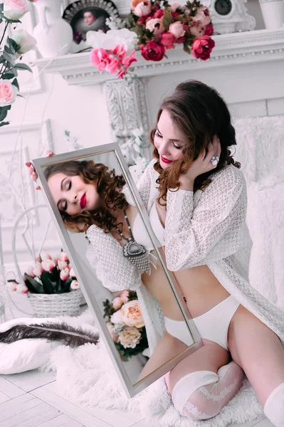 Mooie jonge sexy vrouw, kijken in de spiegel het dragen van witte lingerie, kamer versierd met bloemen. Perfecte make-up. Beauty Beli parfum mode. Wimpers. Studio geretoucheerd schot. — Stockfoto