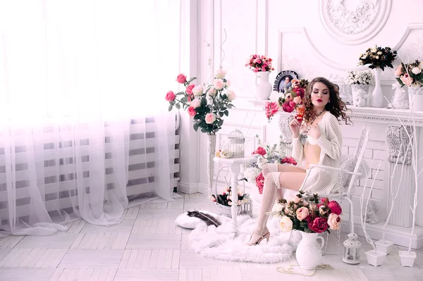 Mulher sexy jovem bonita, segurando um copo e sentado em uma cadeira, vestindo lingerie branca, quarto decorado com flores. Maquiagem perfeita. Moda de beleza. Cílios. Estúdio retocado tiro . — Fotografia de Stock