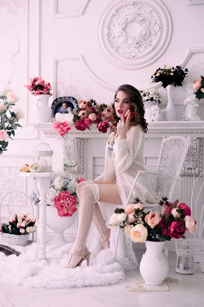 Mulher sexy jovem bonita, segurando um copo e sentado em uma cadeira, vestindo lingerie branca, quarto decorado com flores. Maquiagem perfeita. Moda de beleza. Cílios. Estúdio retocado tiro . — Fotografia de Stock