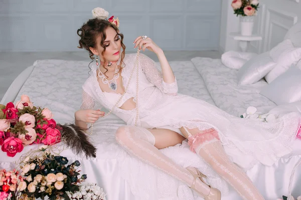 Beautiful young sexy woman sitting on white bed, wearing white lace dress, hair decorated with flowers. Perfect makeup. Beauty fashion. Eyelashes. Studio retouched shot.