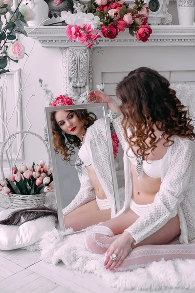 Красивая молодая сексуальная женщина, смотрящая в зеркало в белом нижнем белье, комната украшена цветами. Идеальный макияж. Красота. Ресницы. Ретушированный снимок . — стоковое фото
