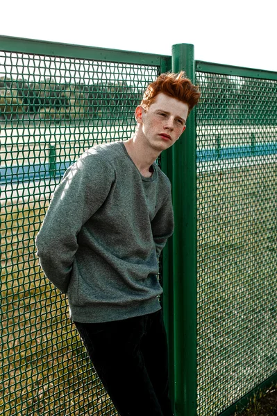 Een knappe red haired modieuze jongen permanent in de buurt van stadion in zonnige dag buiten, het dragen van casual kleding — Stockfoto