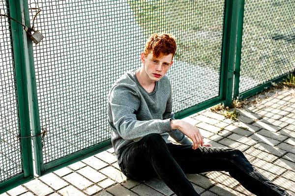 One young handsome red haired fashionable boy sitting near stadium in sunny day outdoor , wearing casual clothing. Close up, retouched shot. — Stock Photo, Image