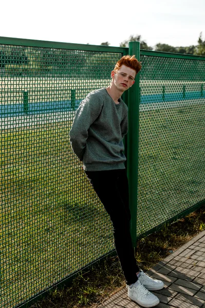 One young handsome red haired fashionable boy standing near stadium in sunny day outdoor , wearing casual clothing — Stock Photo, Image