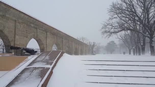 街の公園で冬 吹雪だ 手前は雪に覆われた広い階段 さらに見えるのは石造りのアーケードと古い大砲です アンカーと2つの後退するシルエットが見えます — ストック動画