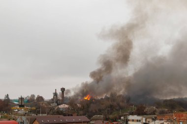 Endüstriyel ya da yarı kentsel bir alanda büyük bir yangın, bulutlu gökyüzüne yoğun, karanlık dumanlar salıyor. Alevler ağaçlar ve binalar arasında görülebilir.