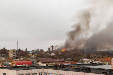 Endüstriyel ya da yarı kentsel bir alanda büyük bir yangın, bulutlu gökyüzüne yoğun, karanlık dumanlar salıyor. Alevler ağaçlar ve binalar arasında görülebilir.