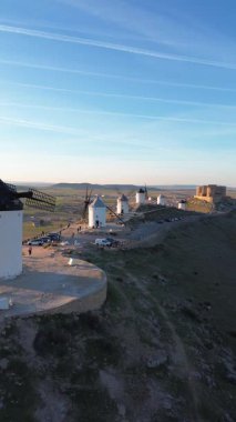 4K dikey hava aracı görüntüleri İspanya, Castilla-La Mancha 'da, Consuegra' da günbatımında geleneksel beyaz yel değirmenlerine doğru ilerliyor. Sıcak altın ışık, manzaralı kırsal ve arka planda dağlar, seyahat için ideal.