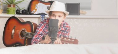 Çocuk video çağrısı, ev müziği dersleri, hobi, vloglama ve yaratılış konseptiyle gitar çalmak için vlog öğreniyor ya da çıkarıyor. Genç gitarist videocu adam oturma odasında gitar çalıyor.
