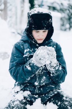 Kulaklık takan sevimli çocuk karda oturup kartopu yapıyor. Gençler iyi eğlenceler ve kış tatillerinin tadını çıkarın. Seçici odak ve tonlama