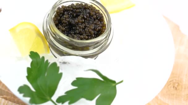 Glass jar with black caviar on ice with lemon, parsley — Stock Video