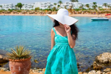 Hasır şapka ve plaj dalgalar üzerinde duran ve uzaklıkta denize bakarak poz zarif elbise giyen genç güzel seksi tabaklanmış esmer kadın