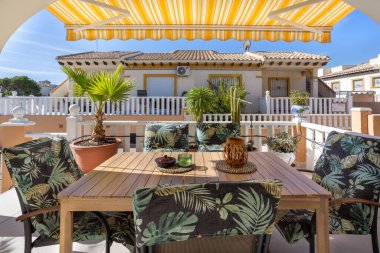 Ahşap bir yemek masası, tropikal yaprak yastıkları, saksı palmiyeleri ve güneşli bir Akdeniz mahallesinde geçen sarı çizgili tentesi olan açık hava verandası..