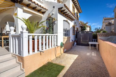 Akdeniz tarzı bir evin yanındaki güneşli bahçe alanında çakıl yolu, bahçe hortumu, küçük depolama kulübesi ve beyaz plastik bir masa var..