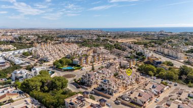 Belirli bir evi vurgulayan açık bir konum işaretine sahip Akdeniz yerleşim bölgesinin havadan görüntüsü, kiremitli çatı, yeşil ağaçlar ve güneşli bir gökyüzünün altındaki uzak deniz manzaralarıyla çevrili..