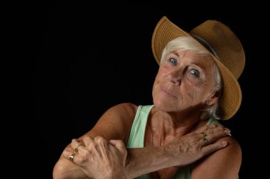 Geniş kenarlı hasır şapka giyen yaşlı bir kadının zarif bir stüdyo portresi. Kollarını nazikçe göğsüne dayar ve sakin ve kendinden emin bir ifadeyle karanlık bir arka plana bakar..