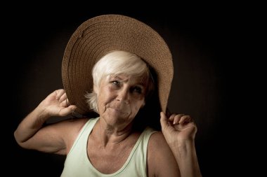 Büyük bir güneş şapkası takan yaşlı bir kadının şık bir stüdyo portresi. Sakin ve kendinden emin bir ifadeyle, şapkasının kenarını hafifçe tutuyor, karanlık bir arka plana karşı cazibe ve zarafet yayıyor..