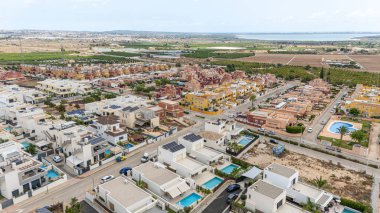 Modern beyaz villalar, havuz ve güneş panelleri, renkli konaklar, yeşil alanlar ve açık bulutlu bir gökyüzü altında uzak kıyı sularının yer aldığı panoramik bir mahalle manzarası..