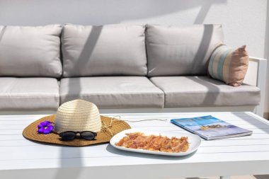 Güneş şapkası, güneş gözlüğü, mavi çiçek, dergi ve açık havada pişirilmiş jambon tabağı olan bir çatı masasının ayrıntıları..