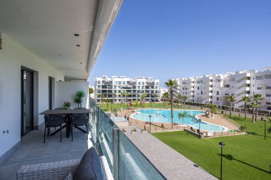 Geniş bir balkon, açık hava yemek alanı geniş bir yüzme havuzu, palmiye ağaçları ve modern beyaz apartman binaları..