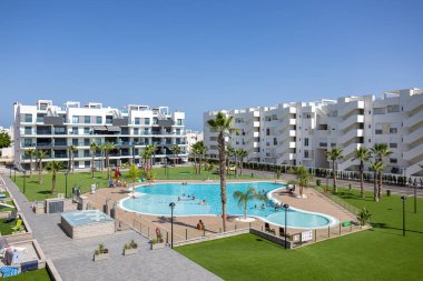 Güneşli bir sitede palmiye ağaçları ve modern beyaz apartmanlarla çevrili geniş bir topluluk havuzu..