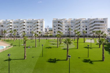 Modern bir sitede yeşil çimenler, palmiye ağaçları ve beyaz apartman binaları olan geniş bir bahçe..