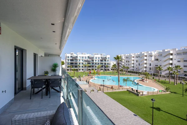 Geniş bir balkon, açık hava yemek alanı geniş bir yüzme havuzu, palmiye ağaçları ve modern beyaz apartman binaları..