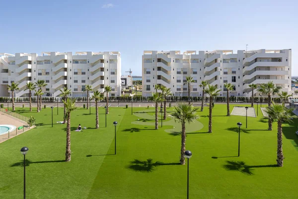 Modern bir sitede yeşil çimenler, palmiye ağaçları ve beyaz apartman binaları olan geniş bir bahçe..