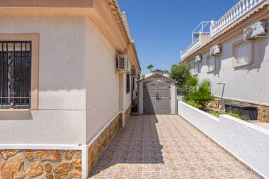 Plastik depolama kulübesi ve güneş ışığında taş kaplı ev duvarlarıyla döşenmiş yan patika.