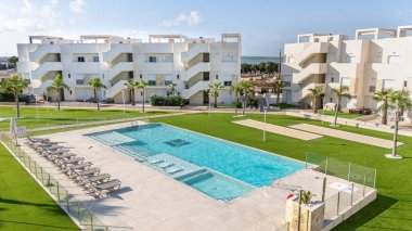 Geniş yüzme havuzu, güneşli yataklar, yeşil peyzaj ve beyaz Akdeniz tarzı binalar..