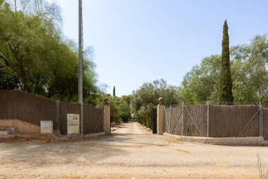 Taştan kapı eşikleri, açık metal kapılar, çakıl yolları ve etrafını çevreleyen ağaçlarla taşra arazisi girişi.
