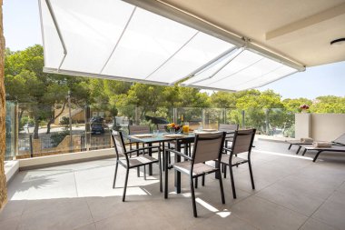 Tente gölgesi, yemek mobilyası, şezlong ve yeşil ağaçların manzarası olan geniş bir teras.