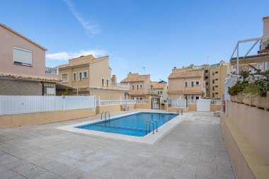 Açık havuz bej evler, döşeme döşeme, çit ve parlak güneşli gökyüzü ile çevrili.