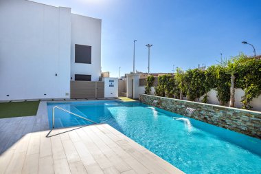 Bitkilerle ve güneşli mavi gökyüzüyle çevrili, taştan şelale özellikli modern havuzu canlandırıyor.