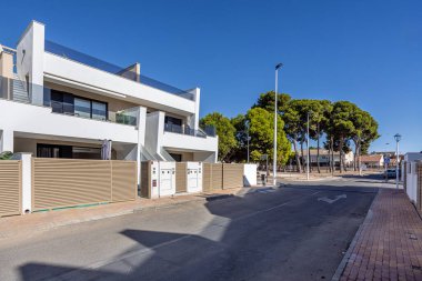 Modern evleri ve etrafındaki çam ağaçlarıyla güneşli bir sokak manzarası.