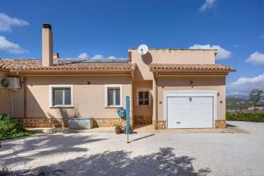 Beige villa exterior with tiled roof, white garage door, gravel driveway, and sunny backdrop.