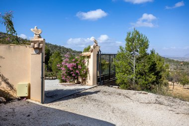 Çiçekler, çam ağaçları, dağ zeminleri ve güneşli bir günde açık gökyüzü ile süslü bir villa kapısı..